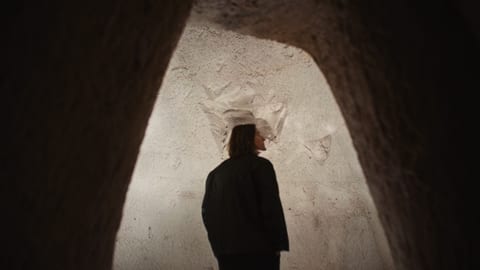 This is a dark image featuring a tunnel with a person standing near the entrance. The tunnel has chalk walls and appears to be illuminated from inside. The l…