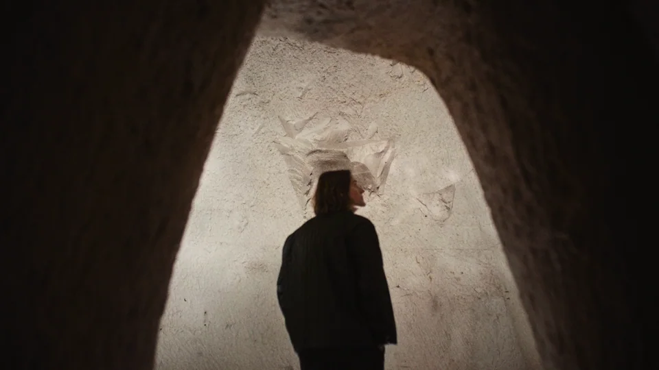 This is a dark image featuring a tunnel with a person standing near the entrance. The tunnel has chalk walls and appears to be illuminated from inside. The l…
