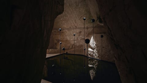 This is a staged photo of an underground tunnel, dark and damp, with water flowing through it. In the foreground, there are floating lanterns that emit a sof…
