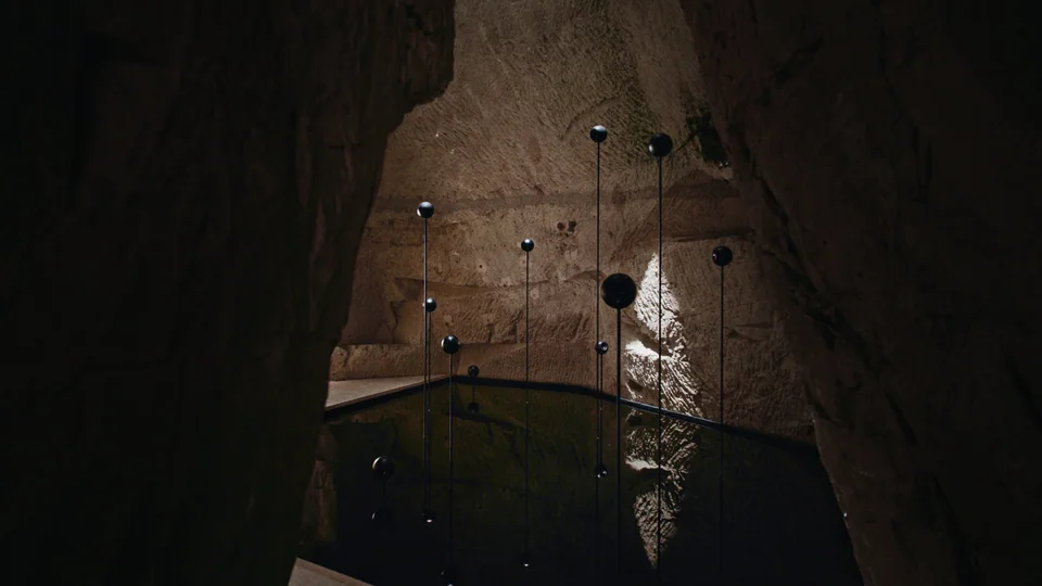 This is a staged photo of an underground tunnel, dark and damp, with water flowing through it. In the foreground, there are floating lanterns that emit a sof…