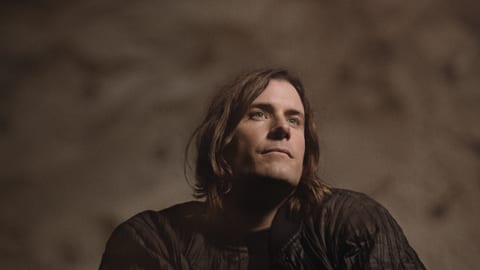 Man in dark room with long hair and beard, contemplating.