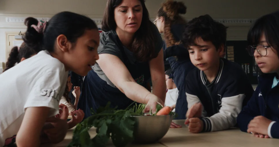 Woman standing behind children at table with vegetables.