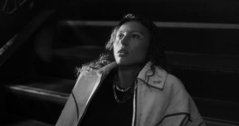 The image shows a woman standing in a room with her head tilted to the side, appearing thoughtful or puzzled. She has light skin and is wearing a dark top wi…