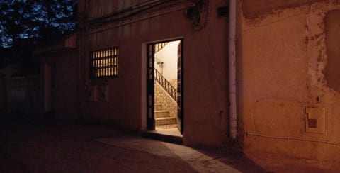 The photo shows a dimly lit doorway at night. There is a single person standing in front of the door. The background is mostly dark, with the exception of th…
